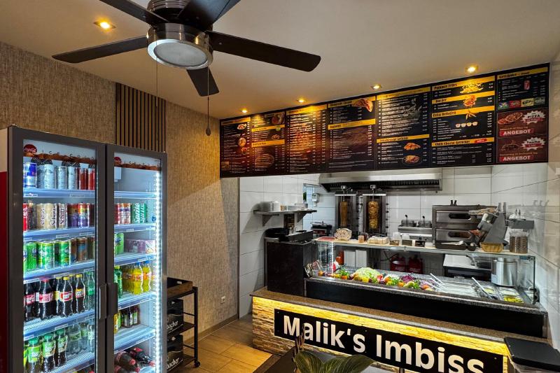 Verkaufstheke mit beleuchtetem Malik’s-Imbiss-Schild, Menütafeln und Getränkekühlschränken